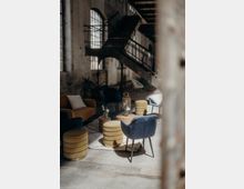 Industrielles Loft mit großen, bogenförmigen Fenstern, groben Backsteinwänden und einer Metalltreppe im Hintergrund. Im Vordergrund steht eine Sitzecke mit modernen, gepolsterten Sesseln, Hockern und kleinen Holztischen, die mit Blumen dekoriert sind.