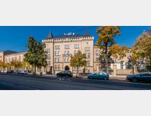 Historisches Backsteingebäude mit symmetrischer Fassade und dekorativen Spitzdächern, umgeben von Bäumen und einem metallenen Zaun; vor dem Gebäude Parkplätze und eine Straßenbahnschiene.