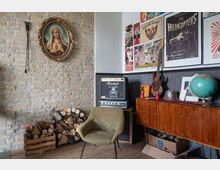 Ein Wohnzimmer mit einer Ziegelwand, an der ein gerahmtes Marienbild hängt. Im Raum befinden sich ein grüner Sessel, gestapeltes Kaminholz, ein Verstärker, eine E-Gitarre an der Wand und eine Holzkommode mit einem Globus und anderen Dekorationsgegenständen; daneben hängen Poster mit Motiven aus der Musik- und Tattoo-Szene.