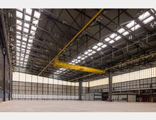 Große, leere Industriehalle mit einem hohen Stahlträgerdach, zahlreichen Oberlichtern und einer auffälligen gelben Laufkranstruktur. Die Wände bestehen aus semi-transparenten Paneelen, die ein gleichmäßiges, diffuses Licht hereinlassen.