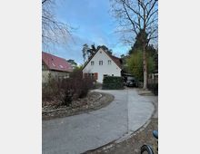 Einfahrt aus Beton zu einem kleinen, weiß verputzten Wohnhaus mit steilem Giebeldach und braunen Fensterläden. Links verläuft ein geschwungener Weg mit niedrigen Sträuchern, rechts stehen ein Zaun und ein Carport neben hohen, teils kahlen Bäumen.