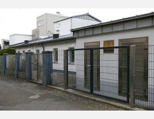 Industriegebäude mit flachem Dach und schlichtem weißem Putz, umgeben von einem Metallzaun mit verschlossenen Toren. An der Fassade befinden sich Warnschilder für Hochspannung.