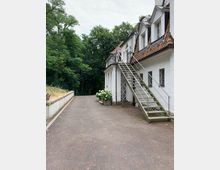 Außenansicht eines weißen Gebäudes mit roten Dachziegeln und mehreren kleinen Fenstern, einer Metallaußentreppe und Blumenkübel mit Hortensien. Im Vordergrund führt ein gepflasterter Weg, umgeben von grünem Wald im Hintergrund.