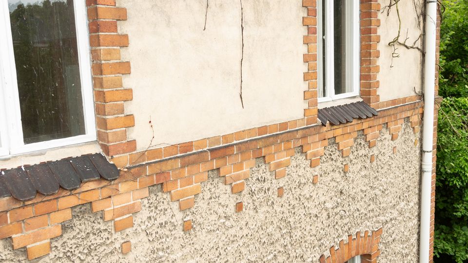 Außenansicht einer älteren Hausfassade mit hellem Putz und sichtbaren Ziegelumrahmungen um die Fenster, teils mit Bogenstürzen. An der Wand sind kletternde Ranken zu sehen, rechts verläuft ein Regenfallrohr, und unten links ragt ein Ziegeldach ins Bild.