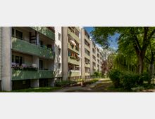 Mehrstöckiges Wohngebäude mit grünen Balkongeländern und dekorierten Balkonen, flankiert von einem schmalen Gehweg mit Grünflächen und großen Bäumen. Im Hintergrund ist ein klarer blauer Himmel zu sehen.