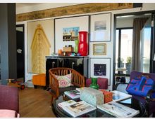 Wohnzimmer mit eklektischem Einrichtungsstil, einschließlich moderner und Retro-Möbel. Der Raum enthält eine schwarze Anrichte, große gerahmte Bilder an der Wand, einen Glastisch mit Büchern und Dekorationen sowie helle Möbel und Accessoires. Große Fenster lassen viel Licht herein.