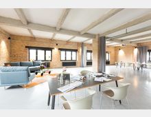 Großzügiger, offener Raum mit Backsteinwänden, freiliegenden Deckenbalken und großen Fenstern. Der Raum ist modern eingerichtet mit einer Sitzecke aus blauen Sofas und mehreren Tischen mit Stühlen, geeignet für Meetings oder Veranstaltungen.