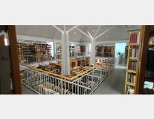 Ein Innenraum einer Bibliothek mit zwei Etagen, Büchersammlungen in Holzregalen und einem zentralen weißen Stützpfeiler. Im Obergeschoss verläuft ein Geländer, das die offene Balustrade umgibt, und durch große Fenster fällt natürliches Licht ein.