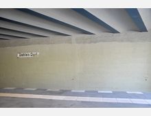 Eine Unterführung mit einer cremefarbenen Betonwand und blauen Deckenbalken. An der Wand ist ein Schild mit der Aufschrift „Dahlem-Dorf“ befestigt, darunter verläuft ein gepflasterter Gehweg in hellen und dunklen Tönen.