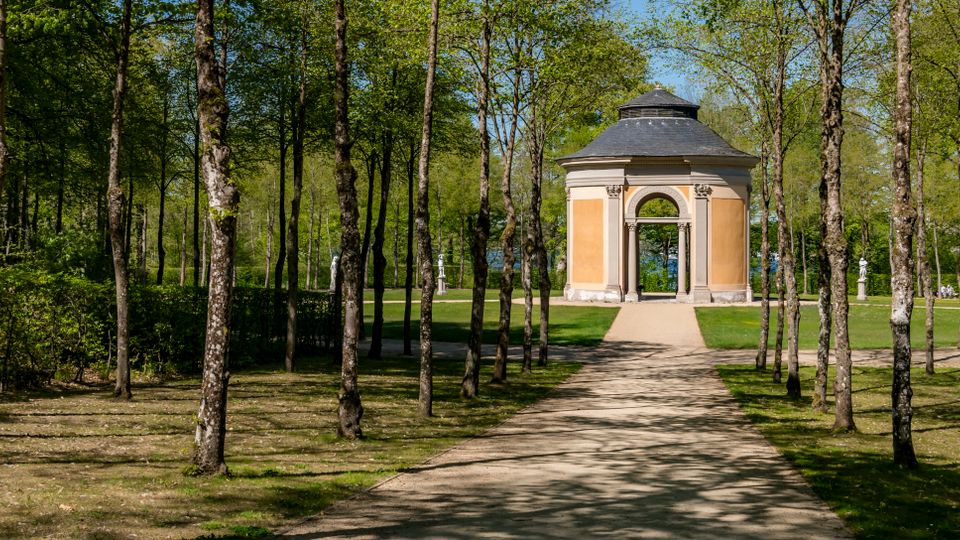 Tree-lined gravel path in a park leading to a small octagonal pavilion with pale orange walls, arched openings, and a dark domed roof. The pavilion sits on a grassy lawn surrounded by tall trees and scattered classical statues.