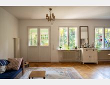 Helles Wohnzimmer mit Fischgrät-Parkett, großem Teppich und Kronleuchter sowie Doppeltür und mehreren Sprossenfenstern. Unter den Fenstern stehen Heizkörper und eine weiße Kommode mit Spiegel; links ist ein Sofa zu sehen.