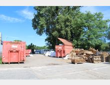 Ein offener Lagerplatz mit roten Containern, gestapelten Paletten und Holzbrettern. Im Hintergrund sind große Bäume sowie gestapelte Metallgitter zu sehen, unter klarem blauem Himmel.