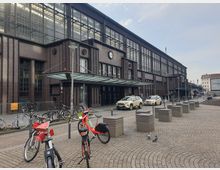 Außenansicht eines historischen Backsteinbahnhofs mit großen Fensterfronten und einem überdachten Eingangsbereich. Im Vordergrund stehen geparkte Fahrräder auf einem gepflasterten Platz, während Autos und Fußgänger den Bereich nutzen.
