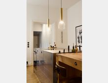 Moderne Küche mit hellem Parkettboden, einer Arbeitsfläche aus Edelstahl und hölzerner Kochinsel. Zwei goldene Hängelampen hängen von der Decke, und an der Wand hängt ein gerahmtes Poster.