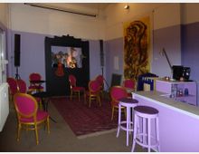 Kleiner Veranstaltungsraum mit lila Wänden, buntem Teppich und runden Tischen mit pink-goldenen Stühlen. Im Hintergrund befinden sich eine Puppentheaterbühne und ein großes Gemälde, rechts eine kleine Bar mit Hockern und Kaffeemaschine.