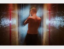 Ein Mann steht mit freiem Oberkörper in einer duschähnlichen Umgebung zwischen roten Backsteinwänden, wobei Wasser in feinem Nebel sprüht und ihn von hinten beleuchtet. Die Szene zeigt ein enges, feucht wirkendes Ambiente mit bunten Lichtreflexionen.