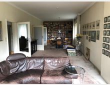 Wohnzimmer mit einem braunen Ledersofa im Vordergrund, einem runden Beistelltisch und einem Teppichboden. Im Hintergrund ein Essbereich mit Regalen voller Bücher, einem großen Tisch mit Stühlen und einer Pendelleuchte; an der rechten Wand sind Bilder und Gemälde aufgehängt.