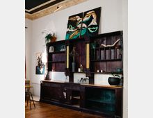 Holzvertäfelter Raum mit einem großen, dunklen Vintage-Regalschrank, dekoriert mit Vasen, Pflanzen und Kerzen. Über dem Schrank hängen zwei abstrakte Gemälde, umgeben von einer hohen Decke mit verzierten Stuckelementen.