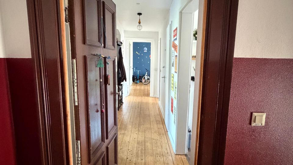 Blick durch einen dunkelbraunen Türrahmen in einen langen, schmalen Flur mit hellem Holzboden und weißen Wänden. Entlang des Gangs befinden sich mehrere Türen, eine Deckenlampe und am Ende eine blau gestrichene Wand mit abgestelltem Fahrrad.