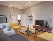 Modernes Wohnzimmer mit hellgrauen Wänden, einer gelben und grauen Sofaecke und einem gläsernen Couchtisch. Auf einem Holzmöbel befindet sich ein Fernseher, daneben steht eine Vase mit Zweigen; im Hintergrund führt eine weiße Tür in einen angrenzenden Raum.