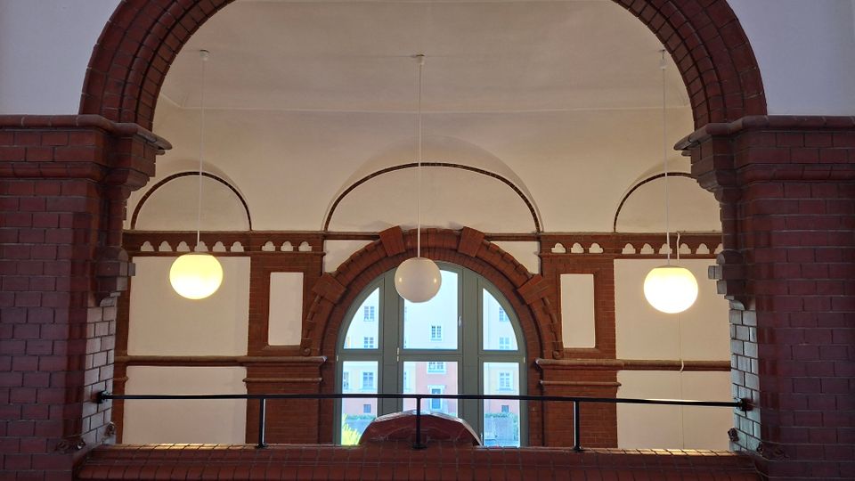 Innenansicht eines Flurs mit großen Backsteinbögen und weiß verputzten Gewölbedecken, beleuchtet von drei runden Hängelampen. In der Mitte befindet sich ein hohes Rundbogenfenster mit Backsteinrahmung und einem Geländer davor; draußen sind Fassaden gegenüberliegender Gebäude zu sehen.