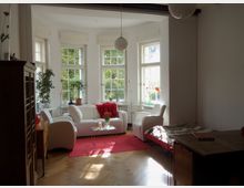 Heller Wohnraum mit großen Fenstern, hellem Holzboden und weißen Möbeln, darunter ein Sofa und zwei Sessel. Eine rote Decke und ein Teppich setzen Akzente, während Pflanzen und Deko auf den Fensterbänken stehen.