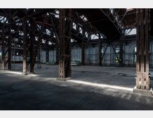 Industriehalle mit hohen Holzträgerkonstruktionen, sichtbaren Stahlverstrebungen und großen Fenstern an den Seitenwänden. Der Betonboden ist teilweise beleuchtet, und in der Halle befinden sich abgedeckte Gegenstände und Gerüste.