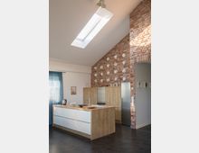 Moderne Küche mit einer freistehenden Kücheninsel aus Holz und weißen Schubladen, einem Metallspülbecken und darauf arrangierten Küchenutensilien. Im Hintergrund eine Backsteinwand mit dekorativen weißen Elementen, ein hoher Kühlschrank aus Edelstahl und ein Oberlicht, das die Szene erhellt.