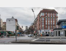 Straßenecke mit einem markanten Gebäude in Backsteinoptik, das eine Filiale der Deutschen Bank beherbergt, sowie einer U-Bahn-Station mit einem Eingang der Linie U4 (Bayerischer Platz). Im Hintergrund ist eine Kirche mit Turmuhr zu erkennen, umgeben von Bäumen und Wohnhäusern.