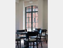 Heller Innenraum eines Cafés oder Restaurants mit dunklen Holztischen und Stühlen, Sitzflächen in hellem Polster. Im Hintergrund ein großes Fenster mit Sprossen, das den Blick auf eine aus rotem Backstein bestehende Außenfassade bietet.