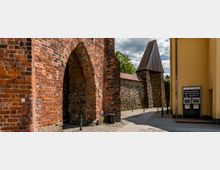 Historisches Stadttor aus rotem Backstein mit spitzem Bogen und angrenzender Steinmauer; im Hintergrund ein kleines Turmdach und grüne Bäume, rechts ein gelbes Gebäude mit Schildern.