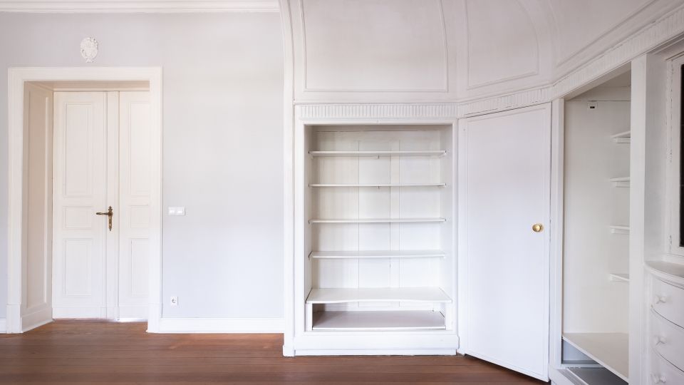 Heller, leerer Innenraum mit weiß gestrichenen Wänden, hoher Decke mit Stuck und dunklem Holzboden. Zu sehen sind eine Tür sowie ein eingebauter Wandschrank mit offenen Regalböden und geöffneter Schranktür.