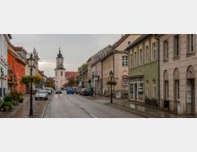 Eine gepflasterte Straße in einer historischen Altstadt mit farbigen Gebäuden auf beiden Seiten, Laternen mit Blumenschmuck und parkenden Autos. Im Hintergrund steht ein Kirchturm mit einer hellen Fassade und dunkler Turmspitze.
