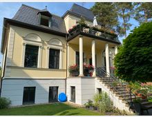 Gelbe Villa mit zwei Stockwerken und einem dunkelgrauen Mansardendach, verziert mit klassischen Elementen wie Säulen, Bögen und Stuckornamenten. Eine Außentreppe führt zu einer überdachten Terrasse mit Blumen, umgeben von einem Garten mit Rasen, Sträuchern und Bäumen.