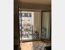 Blick aus einem Wohnzimmer mit Parkettboden und Teppich durch geöffnete doppelte Balkontüren auf einen kleinen Balkon mit Holzmöbeln, einem dekorativen Zickzack-Teppich und einer angrenzenden Wohnumgebung mit gelben Gebäudefassaden.