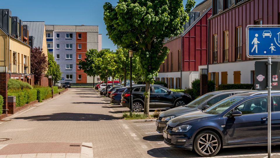 Ruhige Wohnstraße mit gepflasterter Fahrbahn, parkenden Autos entlang der rechten Seite und Bäumen in der Mitte. Links und rechts stehen moderne Reihenhäuser mit farbigen Fassaden und großen Fenstern; im Vordergrund ist ein blaues Verkehrszeichen für einen verkehrsberuhigten Bereich zu sehen.