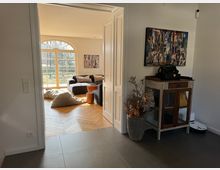 Ansicht eines Wohnbereichs durch eine geöffnete Doppeltür. Der Raum hat helle Wände, einen Holzfußboden in Fischgrätmuster, ein großes Sofa vor bodentiefen Fenstern mit Rundbogenelementen, und Dekorationen wie Kissen und moderne Wandkunst. Im Vordergrund steht ein dekoratives Holzschränkchen mit darauf platzierten Gegenständen.