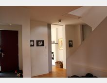 Moderner Eingangsbereich eines Hauses mit weißen Wänden, einer roten Tür und Holzboden. An den Wänden hängen Schwarz-Weiß-Fotografien, und in der Nische im Hintergrund befindet sich ein Korb mit Kaminholz.