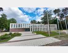 Modernes Rathausgebäude in Dallgow-Döberitz mit einer rechteckigen Architektur, großen vertikalen Fenstern und einer Fassade aus hellem Stein und Ziegel. Vor dem Gebäude befinden sich eine gepflasterte Fläche, Grünflächen und mehrere Bäume.