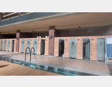 Historische Umkleidekabinen in einem Schwimmbad mit orangefarbenen, gefliesten Wänden und grünlichen Holztüren. Im Vordergrund ist der Rand eines Schwimmbeckens mit einem Metallgeländer zu sehen.