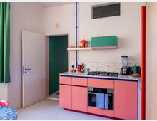 Colorful modern kitchen with pink lower cabinets, a marble-look countertop, and a built-in oven beneath a gas cooktop. White walls feature open shelves and a green wall cabinet, with exposed red and blue pipes, a small high window, and an open door leading to another room.