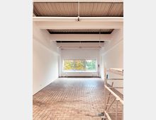 Ein leerer Raum mit hellen Wänden, einem Boden aus beigen Fliesen und einer Decke mit freiliegenden Metallträgern. Am Ende des Raums befindet sich ein großes Fenster mit Blick auf herbstliche Bäume, daneben steht ein Heizkörper. Rechts im Vordergrund ist ein Gerüst sichtbar.