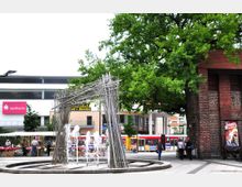 Stadtplatz mit einem modernen Brunnen aus Edelstahlstäben und sprudelnden Fontänen im Vordergrund. Im Hintergrund befinden sich ein historisches Backsteingebäude, Bäume, ein Sparkassengebäude sowie öffentliche Verkehrsmittel und ein Marktstand.