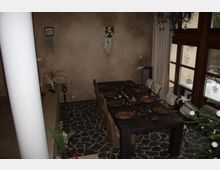 Esszimmer mit dunklem Holztisch, dekoriert für eine Mahlzeit, mit runden Platzdeckchen und dekorativen Elementen. Der Raum hat steinartige Bodenfliesen, eine beige Wand, große Fenster mit Schneeflocken-Aufklebern und einen Weihnachtsbaum im Vordergrund.