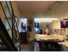 Innenraum eines Ateliers oder Nähstudios mit hohem Raum und industriellem Stil. Zu sehen sind ein großer Arbeitstisch mit Stoffen, Nähmaterialien, Pinnwände mit Bildern und Skizzen sowie Schaufensterpuppen; links führt eine Metalltreppe nach oben, und im Hintergrund hängen Kleidungsstücke auf Stangen.