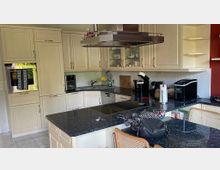 Helle Einbauküche mit cremefarbenen Schränken, schwarzer Granitarbeitsplatte und integrierten Geräten wie einem Backofen und einem Kochfeld. Im Vordergrund steht eine Kücheninsel, und im Hintergrund sind Regale mit Geschirr sowie eine rote Wand sichtbar.