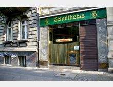 Fassade eines traditionellen Gebäudes mit einer Schultheiss-Werbung über einem Schaufenster. Links davon befinden sich verzierte Glasflächen mit Schriftzügen wie "Bayr. Bier", und an der Hauswand sowie im unteren Bereich sind Graffiti zu sehen.