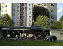 Hochhäuser mit grauer und cremefarbener Fassade umgeben den Eingang der U-Bahn-Station „U-Bhf. Hansaplatz“. Im Vordergrund befinden sich Grünflächen, ein Fahrradstellplatz sowie kleine Geschäfte mit Schildern und Fahrrädern in der Nähe des Eingangs.