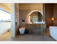 Modernes Badezimmer mit beigefarbenen Wänden und Fliesen. Holzwaschtisch mit rundem, beleuchtetem Spiegel, hängender Designlampe, freistehender Badewanne und Wandtoilette; im Hintergrund eine Heizkörperleiter und eine Holzvertäfelung.