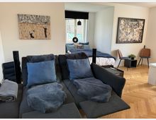 Wohnzimmer mit offener Schlafbereichs-Ecke, ausgestattet mit einer grauen Sofalandschaft, blauen Kissen und Decken. Im Hintergrund ein Bett mit dunkelblauer Bettwäsche, einen Holzboden in Fischgrätmuster, zwei Stühle, ein kleiner Tisch mit Pflanze und Wanddekoration.